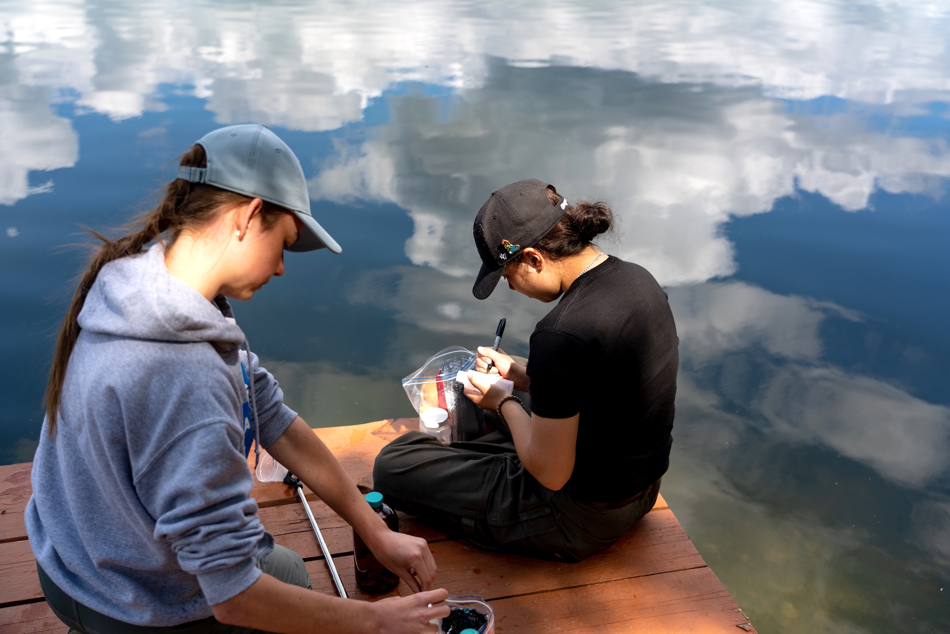 Two young women sitting on a dock by a lake writing labels on empty containters